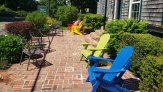 brick laid patio with brightly colored adirondack chairs  facing  the street by wood how with surrounding busheswith