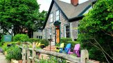 view from street facing the brown house with teal trim and red brick chimney with  Snug Cottage sign, split rail fence and bright adirondack chairs
